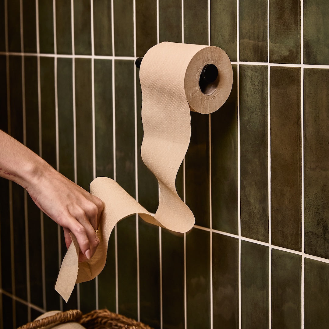 Roll of Naked Paper toilet roll on holder in bathroom with green tiles. A hand pulls some toilet paper from the roll.
